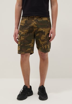 Hombre que lleva pantalones cortos cargo camuflados hasta la rodilla, zapatillas negras y una camiseta negra, de pie frente a un fondo liso.