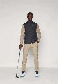 Gilet sans manches bleu marine sur une chemise à manches longues beige, associé à un pantalon beige et des chaussures de golf blanches avec des accents bleus, tenant un club de golf.