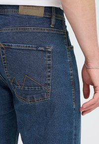 Temno modri jeans iz denima z edinstvenim cikcakovim šivom na zadnjem žepu, s kontrastno oranžno nitjo in teksturirano obdelavo.