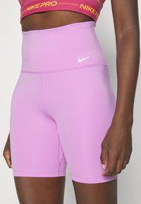 Vysoké sportovní šortky v světlé růžové barvě, vyrobené z hladké látky, s logem Nike v bílé barvě. Obsahují přiléhavý střih a ploché švy.