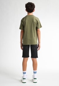 Camiseta de manga corta verde oliva combinada con pantalones cortos negros hasta la rodilla. Zapatillas blancas con detalles en verde y calcetines a rayas azules.