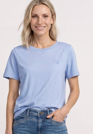Smilende kvinde med blondt hår iført en lyseblå t-shirt med et lille skalsymbol og blå jeans, hånd i lommen.