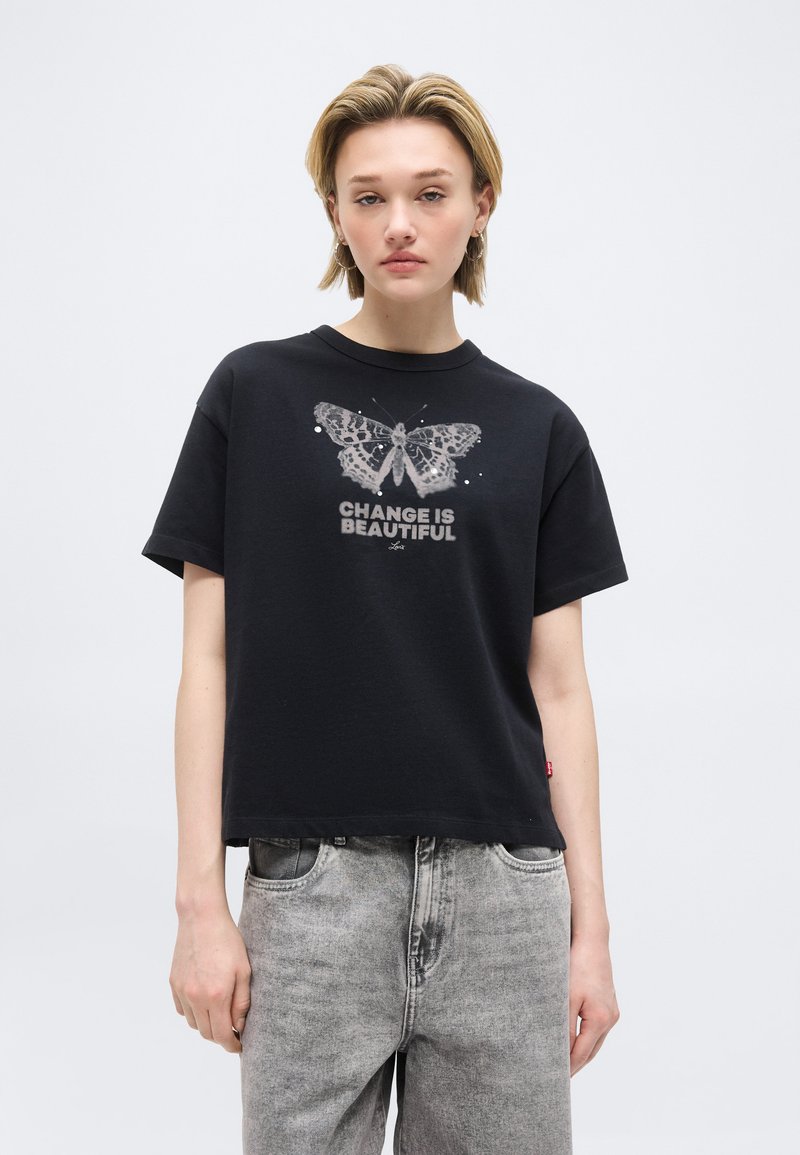 Jeune personne portant un t-shirt noir avec un motif de papillon et le texte « LE CHANGEMENT EST BEAUTÉ », associé à un jean gris délavé sur un fond uni.
