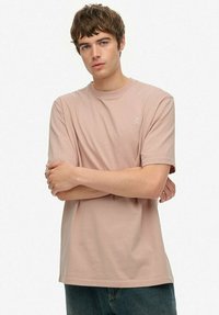 Ung mand med kort brunt hår, der bærer en ensfarvet beige T-shirt og blå jeans, står med armene krydset mod en hvid baggrund.