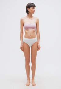 Γυναίκα με κοντά σκούρα μαλλιά, όρθια με γυμνά πόδια, μπροστά σε λευκό φόντο, φοράει ανοιχτό μωβ μπραλέ και λευκά εσώρουχα με ψηλή μέση, κοιτάζει δεξιά.