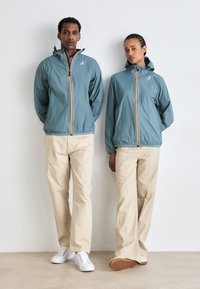 Rompevientos azul claro con capuchas, con franjas amarillas y rojas en la parte delantera, combinados con pantalones beiges y zapatillas blancas. Tela suave y ligera.