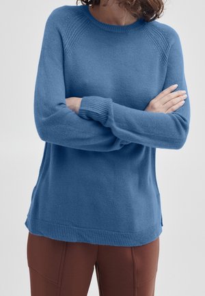 Femme portant un pull en maille bleu à manches longues croisées sur la poitrine et un pantalon marron sur un fond uni.