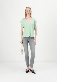 Lys grønn kortermet topp med V-hals, paret med grå tettsittende jeans og svarte spisse hæler, holder en liten hvit veske.