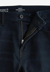 Vaqueros skinny fit en denim índigo oscuro, con cierre de cremallera metálica y un botón. La etiqueta indica "Comfort Stretch" y "Next Performance."