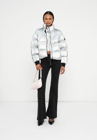 Chaqueta de puffer plateada con diseño cropped y cuello alto, combinada con pantalones negros de campana. La modelo sostiene un pequeño bolso rosa.