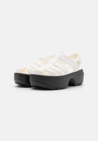 White open-toe clogs with a black platform sole, featuring horizontal and vertical strap designs, made of flexible material.