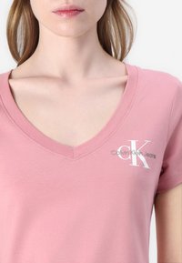 Rozā V-veida krekls, izgatavots no mīksta auduma. Uz kreisā krūts ir baltais "Calvin Klein Jeans" logo, tam ir īsas piedurknes un gluda tekstūra.