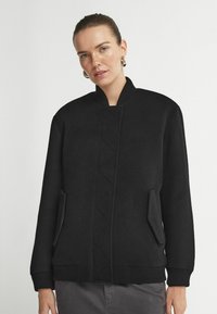 Svart bomberjacka i texturerad kvalitet. Ribbat manschetter och krage. Sidofickor med snedställda öppningar. Har ett centralt diamantmönster.