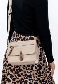 Frau trägt ein schwarzes, geripptes Oberteil und einen Rock mit Leopardenmuster, dazu eine beige Umhängetasche mit Reißverschluss und Schnallendetails.