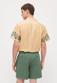 Camicia a maniche corte color beige chiaro con ricami a spirale verde sulle maniche, abbinata a pantaloni corti a quadri verdi e gialli.