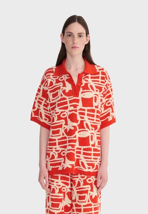 Femme aux longs cheveux bruns portant un polo à manches courtes boutonné, à motif abstrait rouge et crème, avec un pantalon assorti.