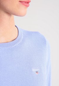 Jasnoniebieski dzianinowy sweter z teksturowym wzorem i ściągaczem przy dekolcie, z małym haftowanym logo na piersi.