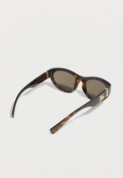 Γυαλιά ηλίου με φαρδιές βραχίονες σε σχέδιο χελωνόκρανου, με σκουρόχρωμους φακούς και λεπτομέρειες λογότυπου σε χρυσή απόχρωση, εμφανιζόμενα από πλευρική και οπίσθια γωνία σε λευκό φόντο.