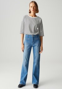 Gestreepte zwart-witte top met brede mouwen, gecombineerd met blauwe flared jeans met voorzakken. Model draagt zwarte schoenen.