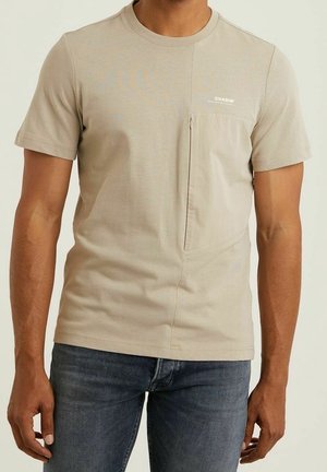Homme portant un t-shirt beige à manches courtes avec une poche poitrine zippée et un petit logo blanc, assorti à un jean bleu.