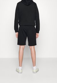 Felpa nera con cappuccio e pantaloni corti abbinati, in cotone misto, vestibilità rilassata, indossati con scarpe da ginnastica verde chiaro e calzini bianchi alla caviglia.