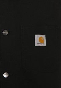 Tessuto nero con una tasca cucita che presenta un'etichetta bianca Carhartt e due bottoni argentati incisi con la scritta "carhartt."