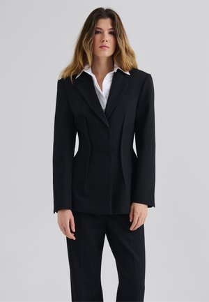 Black tailored blazer with a structured design, sharp notched lapels, and subtle front pleats, paired with a white collared shirt underneath.