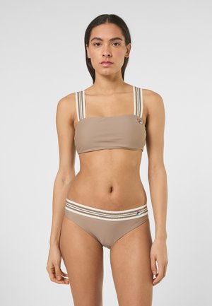 Kvinde stående forfra iført beige badetøj i to dele med elastiske bånd i hvidt og brunt stribet mønster samt små logo-patches.