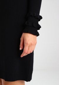 Abito nero in maglia con maniche a volant. Presenta un design aderente e una texture liscia, mettendo in risalto un colore scuro uniforme senza motivi.