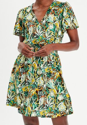 Femme portant une robe à manches courtes et col en V avec un motif floral multicolore, mains jointes à la taille, debout devant un fond uni.