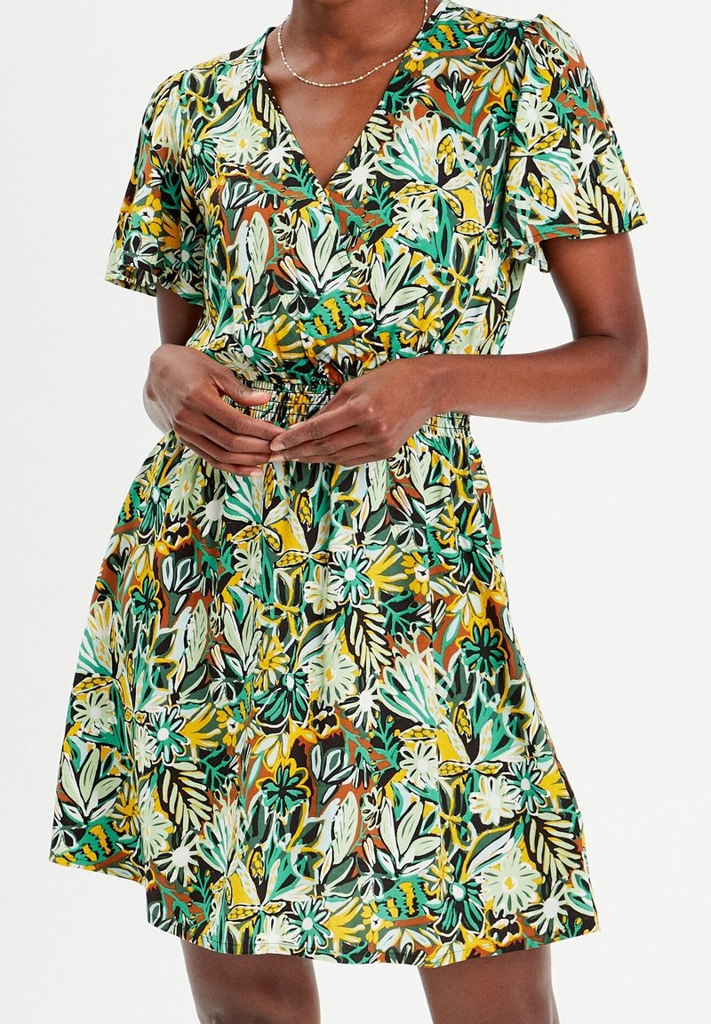 Femme portant une robe à manches courtes et col en V avec un motif floral multicolore, mains jointes à la taille, debout devant un fond uni.