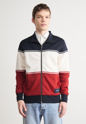 Giacca a zip rossa, crema e blu navy con strisce orizzontali; tessuto morbido; polsini e orlo a costine; etichetta del logo vicino alla parte inferiore sinistra.