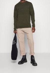 Dunkelgrüner, strukturierter Strickpullover, beiger Hose mit hochgerollten Ärmeln, schwarze Knöchelstiefel und eine schwarze Tragetasche aus Stoff.