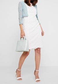 Witte kanten jurk, lichtblauwe kanten cardigan, mintgroene handtas met textuur en witte hakken. De outfit heeft verfijnde patronen en contrasterende kleuren.