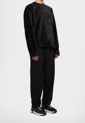 Person trägt einen schwarzen, flauschigen Langarm-Versace Jeans Couture-Pullover, lockere schwarze Hose und schwarz-weiße Turnschuhe.