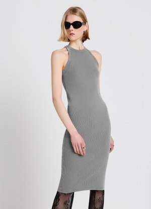 Femme portant une robe midi grise ajustée sans manches, des collants en dentelle noire, de grandes lunettes de soleil noires et des bagues argentées sur un fond clair uni.