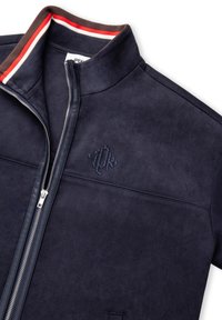 Chaqueta de ante azul marino con un alto cuello, cremallera plateada y logotipo bordado. Presenta acentos a rayas contrastantes en rojo y marrón.