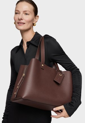 Mujer con una camiseta negra de manga larga que sostiene un gran bolso tote de cuero marrón con herrajes dorados y dos asas para el hombro.