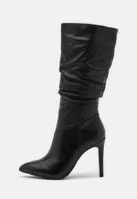 Bota de cuero negro de tacón alto con punta afilada, diseño de parte superior caída y textura suave. Presenta un tacón de aguja y una forma estilizada.