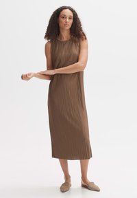 Faltendes, ärmelloses braunes Midi-Kleid mit rundem Ausschnitt. Verfügt über seitliche Schlitze. Getragen mit hellbraunen Slippern.