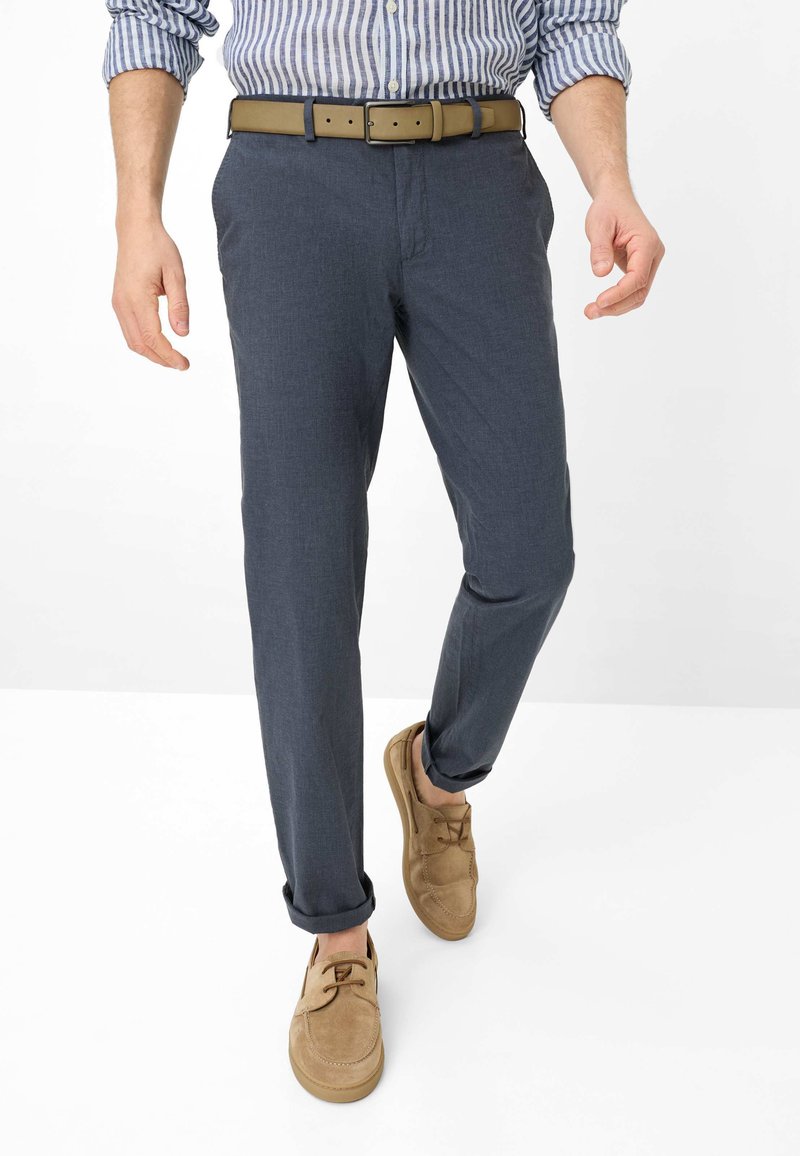 Man loopt en draagt blauwe chino's, een gestreept overhemd in zijn broek gestopt, een tan riem en tan suède casual schoenen tegen een witte achtergrond.