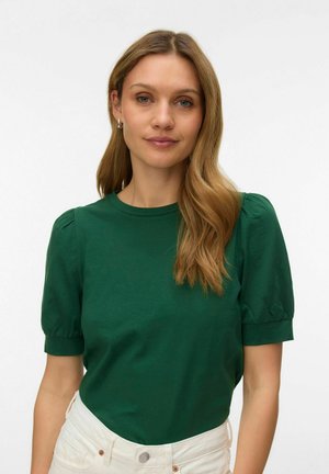 Vrouw met lang blond haar, gekleed in een groen topje met pofmouwen en een witte broek, die recht vooruit kijkt met een neutrale uitdrukking tegen een witte achtergrond.