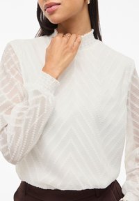 Weiße Bluse mit Zickzackmuster aus durchscheinendem Stoff, mit hohem Kragen und puffärmel, ausgestattet mit gerafften Manschetten und Details.