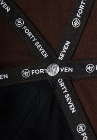Brunt stoff med svarte detaljer, med sammenvevde etiketter som leser "FORTY SEVEN" og en metallknapp-logo i sentrum.