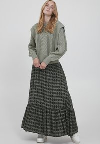 Dick gestrickter, hellgrüner Pullover mit hohem Kragen, kombiniert mit einem langen schwarzen Rock mit geometrischem Muster und Rüschensaum. Weiße Sneakers.
