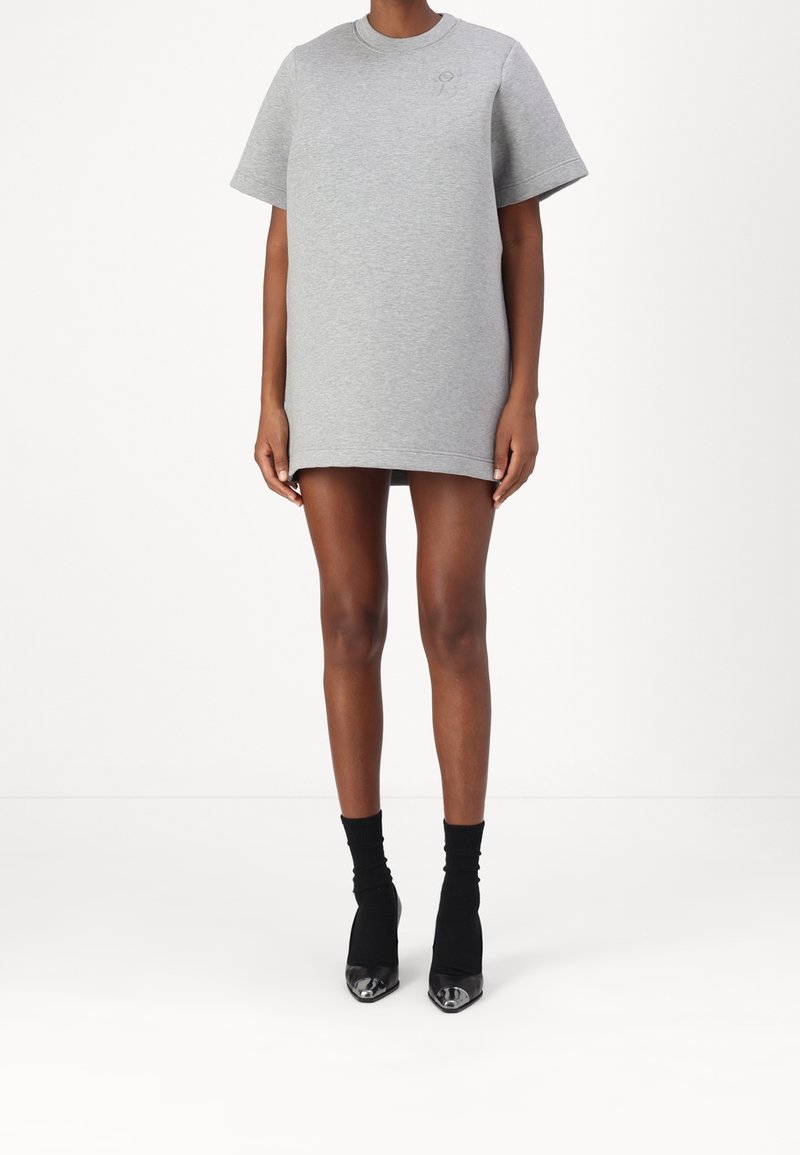 Personne portant une robe t-shirt courte oversize gris clair associée à des bottines chaussettes noires avec des bouts métalliques pointus, debout devant un fond blanc.