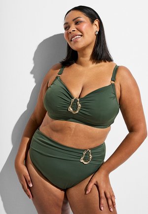 Lachende vrouw gekleed in een olijfgroene, hooggetailleerde bikini met gouden sierknopen, staand tegen een witte muur met schaduw.