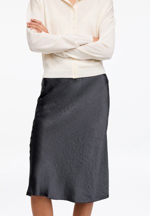 Personne portant un cardigan crème boutonné avec les manches baissées et les bras croisés, associé à une jupe midi noire texturée.