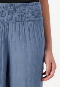 Ljusblå byxor med smockad midja och texturerad väv med vertikala veck, visade tillsammans med en delvis synlig hand.