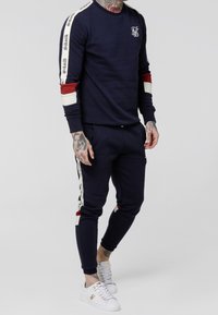 Námořnický modrý set mikiny a tepláků s kontrastním pruhovým designem, žebrovanými manžetami, měkkou tkaninou a bílými teniskami s metalickými akcenty.